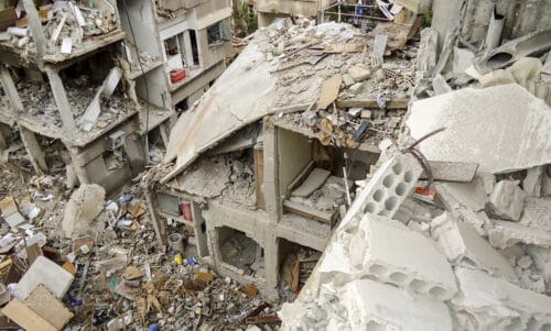 Buildings damaged with lots of trash, white bricks with holes in them, ashes, and scraps of wood and boxes.