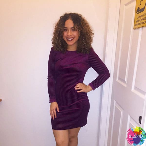 An understanding young woman in dark purple long sleeved corduroy dress stands in front of a white door and wall smiles with her hand on her hip.