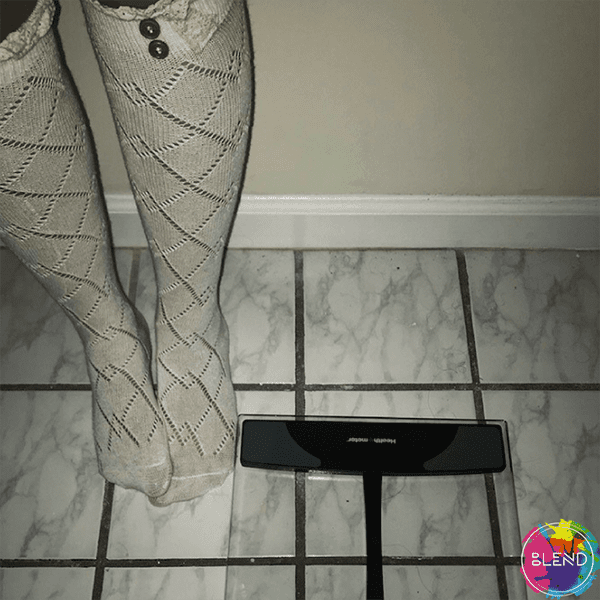A young lady struggling with anorexia nervosa stands next to digital glass scale wearing white socks