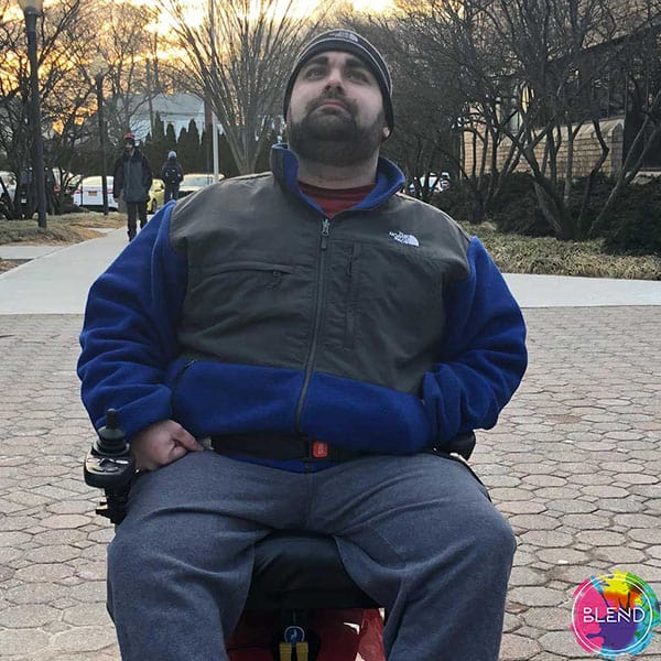 A tall and overweight man with cerebral palsy is wearing a heavy dark blue jacket, while sitting and facing the camera.