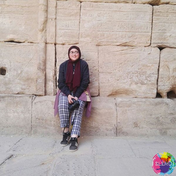 A lady wears a red scarf wrapped around her head, glasses, a long sleeved grey shirt, and grey gingham pants sits on ledge of a large stone wall.