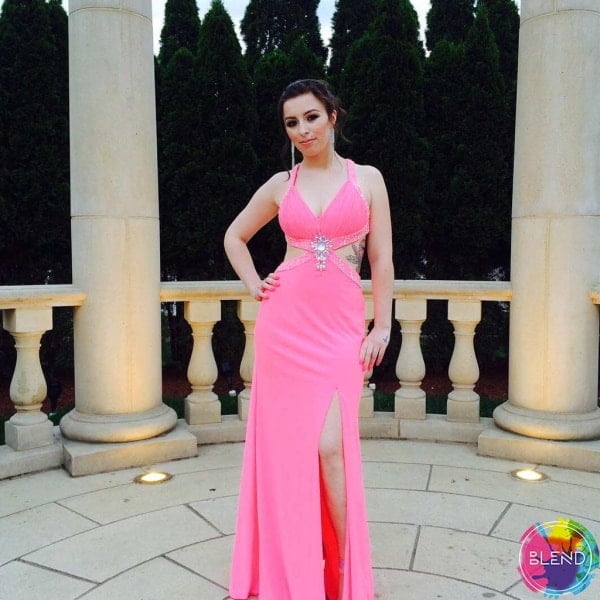 A tall young lady with dark hair wearing a pink dress stands outside of a pillared area.