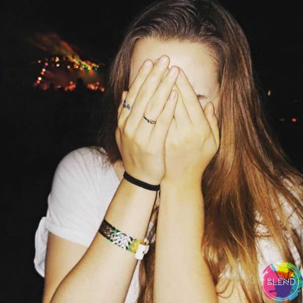 Young lady with brown hair wears a black hairband and multi colored bracelet on her wrist and two rings on her right hand, covers her face with hands.