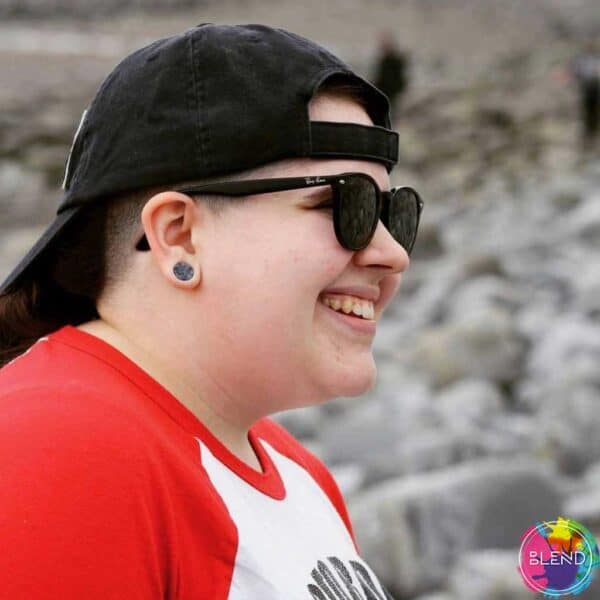 Right side view of gender-queer person with shaved hair black hat on backwards black sunglasses and grey gauge in right ear looks forward and smiles.