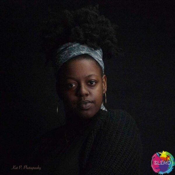 Young black woman with her hair in an updo, a scarf covers up part of her head, wears dangling earrings, looks seriously in the camera in a black room.
