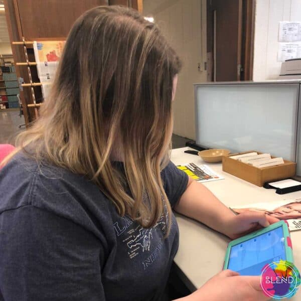 Girl with brown hair and dark grey shirt sitting at a table using her iPad.