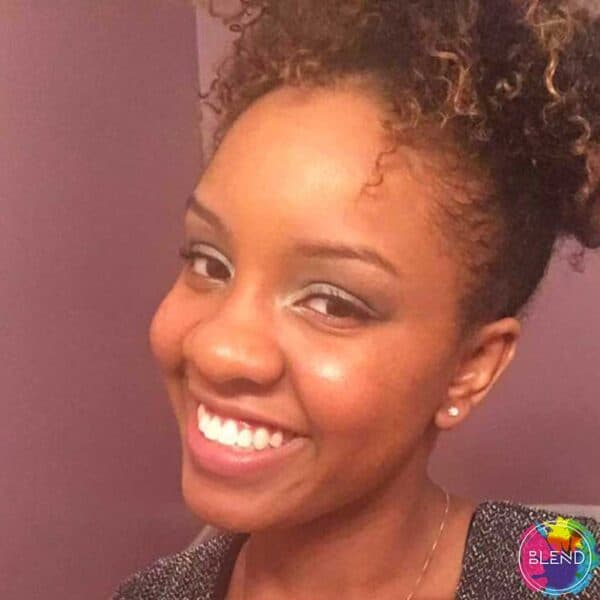 Young woman with rosy cheeks and curly hair in an updo smiles about her journey with white teeth in front of a purple wall.