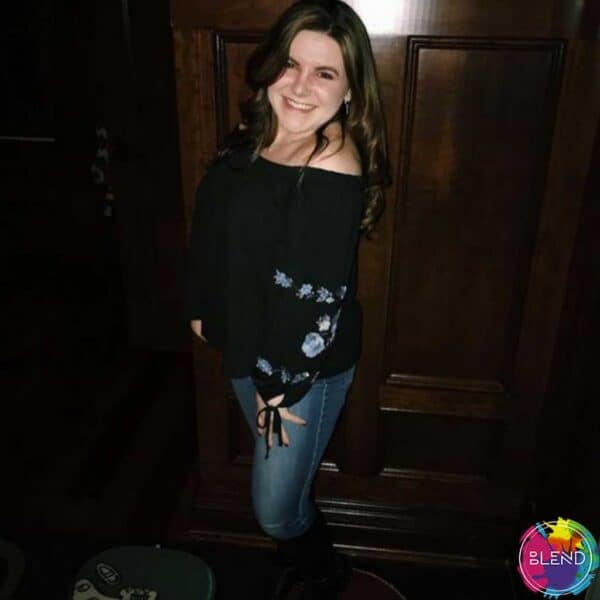 A girl with dark hair, black shit and blue jeans smiling in front of a door.