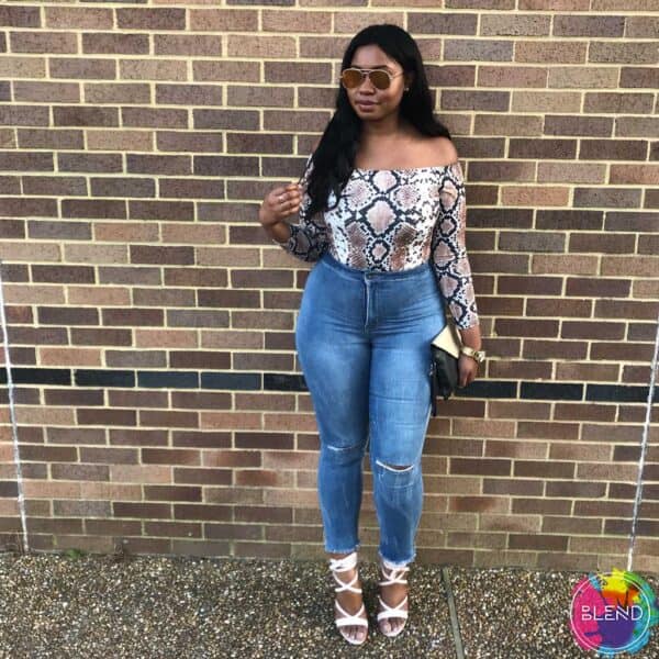 A black girl with dark hair and sunglasses wearing a white shirt and blue jeans standing in front of a brick wall