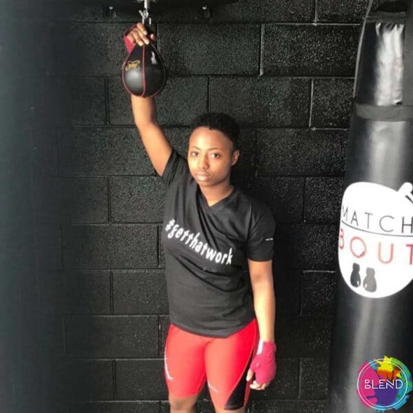 A black girl with dark hair wearing a black shirt, red shorts, and pink gloves standing in front a black wall.