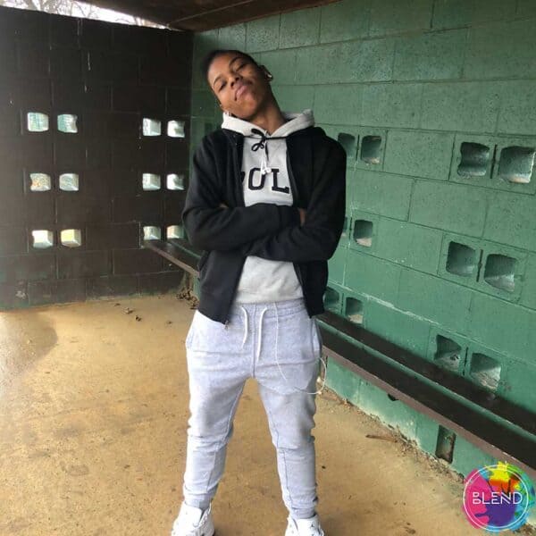 young black guy in grey sweat pants, white tennis shoes and hoodie with black jacket over it, stands with arms crossed head cocked, smirking in a batting cage.