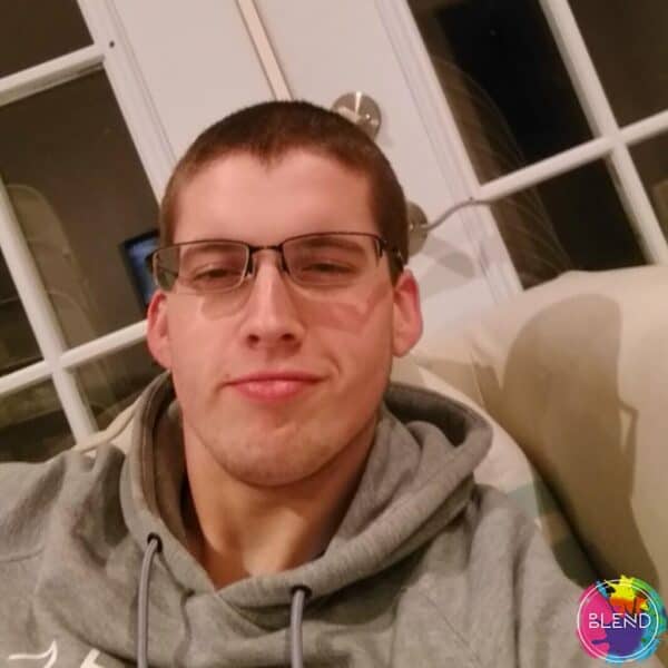 A man with buzzed brown hair, glasses and gray sweatshirt, sitting on a couch smiling at the camera.
