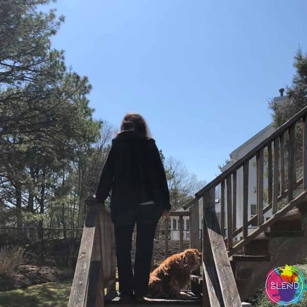 A woman with dark hair, wearing a black jacket standing on a wooden staircase in her backyard with her dog.
