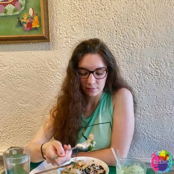 A girl with long brown hair and glasses wearing a cyan shirt while eating food.