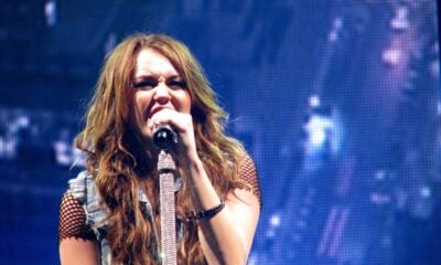 Miley Cyrus with brown hair singing in a microphone at a music concert.