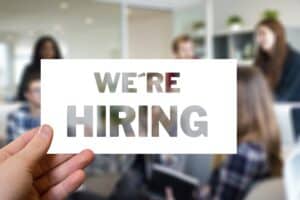 cover Sign being held up in an office, in front of a group of people saying "We're Hiring"