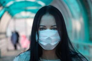 A dark-haired young lady wearing a blue sweater, and a white mask indoors.