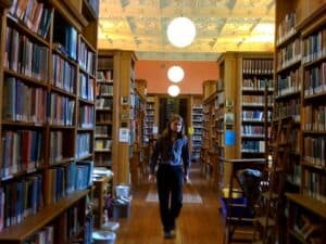 cover Person in a blue sweater and dark pants walking in between rows of book shelves in a library towards the camera