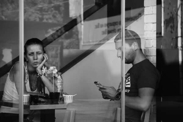 A man and a woman presumably at a coffee shop, the woman looking sad towards the camera behind the window pane, and the man focused on his phone texting