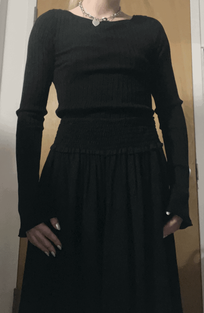 A woman wearing a black dress and a black sweater stands confidently, showcasing her elegant style.
