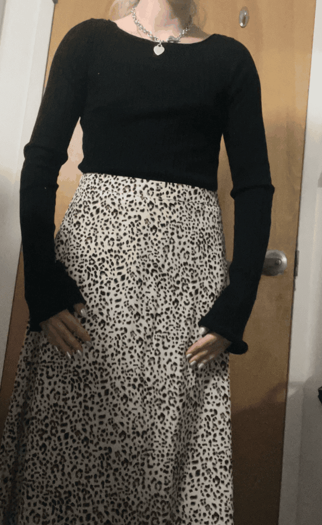 A woman wearing a leopard print skirt and a black sweater stands confidently, showcasing her stylish outfit.