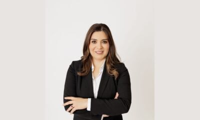 Professional portrait of a woman with long brown hair wearing a black suit and white shirt, smiling with her arms crossed.