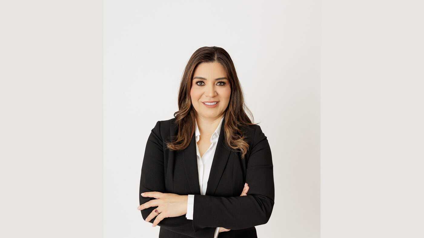 Professional portrait of a woman with long brown hair wearing a black suit and white shirt, smiling with her arms crossed.