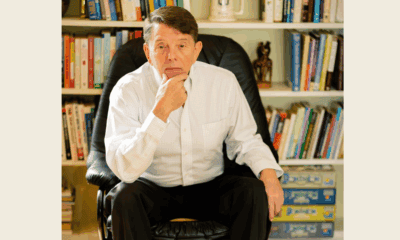 Middle-aged man in a white collared shirt sits in a black leather chair with bookshelves filled with books and games behind him.