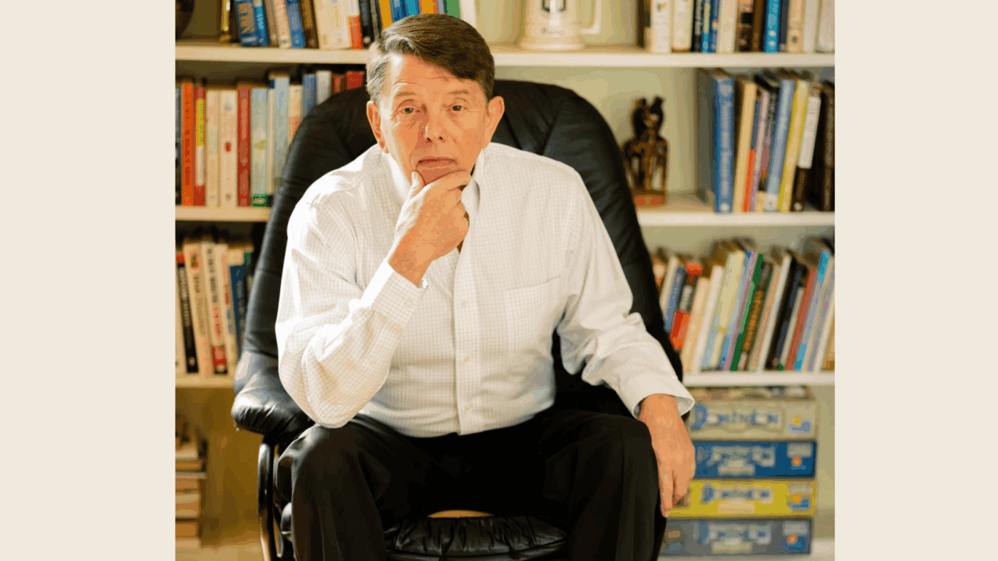 Middle-aged man in a white collared shirt sits in a black leather chair with bookshelves filled with books and games behind him.
